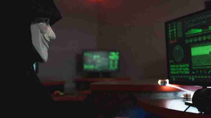 A person wearing a hacker mask operates a computer in a dimly lit room with digital displays., tags: auf den von - pexel